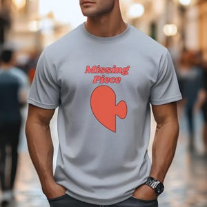 Puede incluir: Una camiseta gris con el texto "Missing Piece" en rosa y una pieza de rompecabezas en forma de corazón rosa.