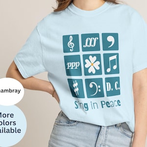 May include: A light blue short-sleeve t-shirt featuring a grid of nine teal squares containing white musical symbols, including a treble clef, a flower, and the text D.C. Below the grid, the text Sing in Peace is printed in teal lettering.