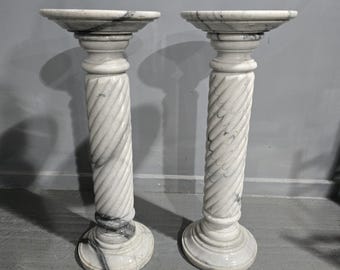 May include: Two white marble pedestals with a spiral column design. The pedestals have a round base and a circular top. The marble has gray veining throughout. The pedestals are suitable for displaying sculptures or plants.