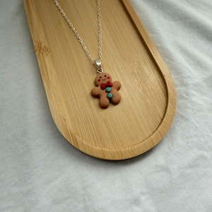 Puede incluir: Collar de cadena plateada con un colgante de hombre de jengibre. El colgante es marrón con una pajarita roja y dos botones turquesa. El collar se muestra en una bandeja de madera.