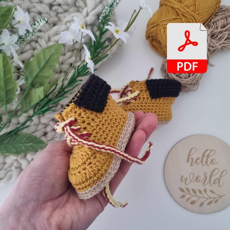 Crocheted baby booties in yellow and brown with red laces. The booties are shaped like hiking boots and have a black fabric detail on the top.