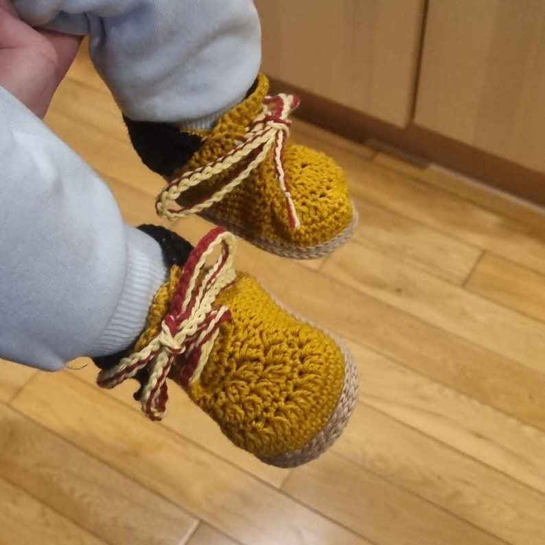 A pair of crocheted baby booties in a mustard yellow colour with a red and white ribbon detail. The booties have a crocheted sole.