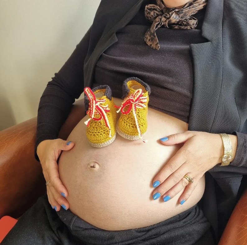 A pregnant person with a large belly, wearing a black blazer and blue jeans, with two small yellow and red crocheted baby shoes resting on their stomach.