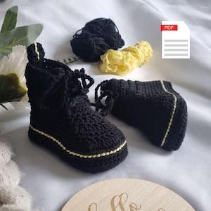 Może przedstawiać: Para czarnych, szydełkowych bucików dla niemowląt z żółtym wykończeniem. Buciki mają sznurowany wzór i są wykonane z fakturowanym ściegiem.