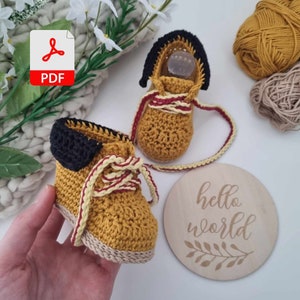 A hand holding a pair of crocheted baby booties in yellow and brown with red and white laces. The booties are on a white surface next to a wooden sign that says "hello world".