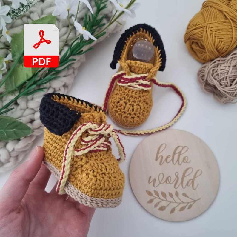 A hand holding a pair of crocheted baby booties in yellow and brown with red and white laces. The booties are on a white surface next to a wooden sign that says "hello world".