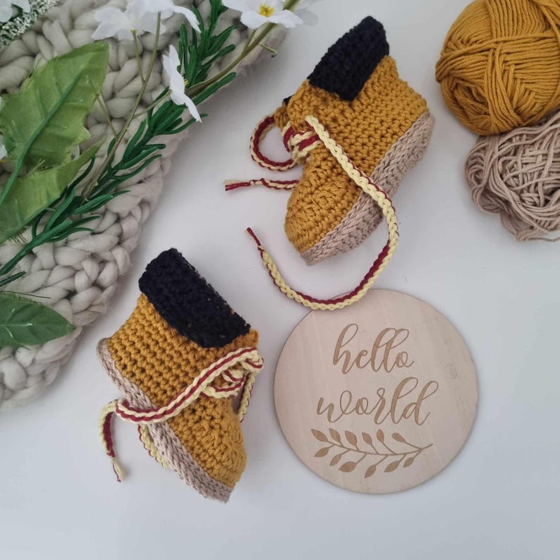 A pair of crocheted baby booties in yellow and brown with red and white laces. The booties are on a white surface next to a wooden sign that says "hello world".