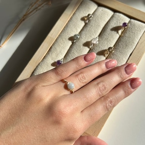Op de afbeelding: Een hand toont een goudkleurige, met draad omwikkelde ring met een ronde, lichtgekleurde edelsteen. Andere ringen met verschillende edelstenen worden getoond in een houten sieradendoos. De ringen liggen op een beige stof.