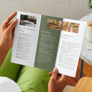 Op de afbeelding: Een persoon houdt een brochure vast met tekst over kerncentrering, ademhaling en uitlijning. De brochure bevat afbeeldingen van yogahoudingen. Een witte mok met koffie staat op een tafel.