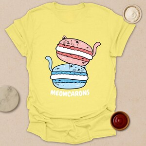 May include: Yellow t-shirt featuring a cartoon illustration of two cat-shaped macarons, one pink and one blue, stacked on top of each other. The text "MEOWCARONS" is printed below the macarons.