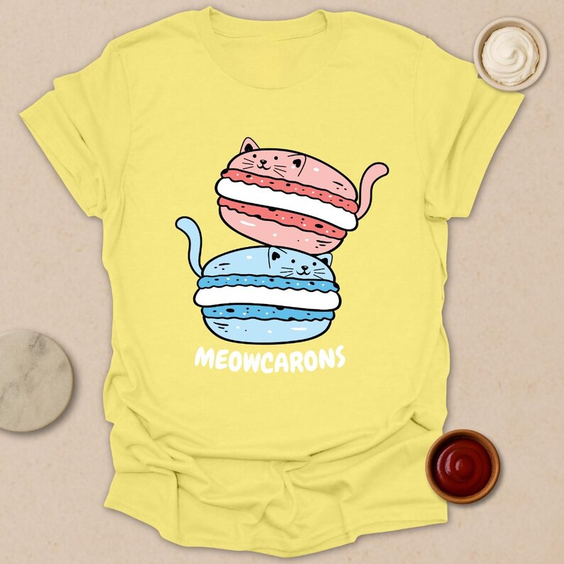 May include: Yellow t-shirt featuring a cartoon illustration of two cat-shaped macarons, one pink and one blue, stacked on top of each other. The text "MEOWCARONS" is printed below the macarons.