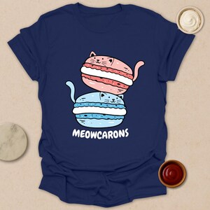 May include: Navy blue t-shirt featuring a playful illustration of two cat-shaped macarons, one pink and one blue, stacked on top of each other. The text "MEOWCARONS" is printed below the macarons.