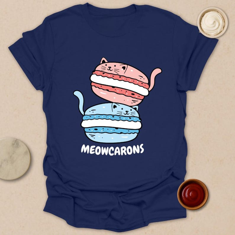 May include: Navy blue t-shirt featuring a playful illustration of two cat-shaped macarons, one pink and one blue, stacked on top of each other. The text "MEOWCARONS" is printed below the macarons.
