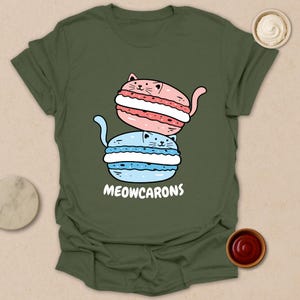 May include: Olive green t-shirt featuring a playful graphic of two cat-shaped macarons, one pink and one blue, stacked on top of each other. The text "MEOWCARONS" is printed below the macarons in white. The shirt is a casual wear item.