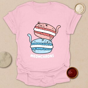 May include: A light pink t-shirt featuring a stacked illustration of two cat-shaped macarons. The top macaron is pink with a red filling, and the bottom one is blue with a white filling. The text "MEOWCARONS" is printed below the macarons.