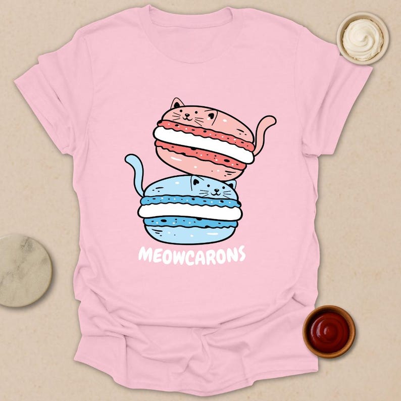 May include: A light pink t-shirt featuring a stacked illustration of two cat-shaped macarons. The top macaron is pink with a red filling, and the bottom one is blue with a white filling. The text "MEOWCARONS" is printed below the macarons.
