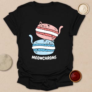 May include: Black t-shirt featuring a whimsical design of two stacked cat-shaped macarons. The top macaron is pink with white filling, and the bottom is blue with white filling. The text "MEOWCARONS" is printed below the macarons.