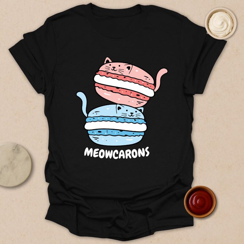 May include: Black t-shirt featuring a whimsical design of two stacked cat-shaped macarons. The top macaron is pink with white filling, and the bottom is blue with white filling. The text "MEOWCARONS" is printed below the macarons.