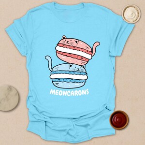 May include: Light blue t-shirt featuring a cartoon illustration of two cat-shaped macarons, one pink and one blue, stacked on top of each other. The text "MEOWCARONS" is printed below the macarons. The shirt is a casual wear item.
