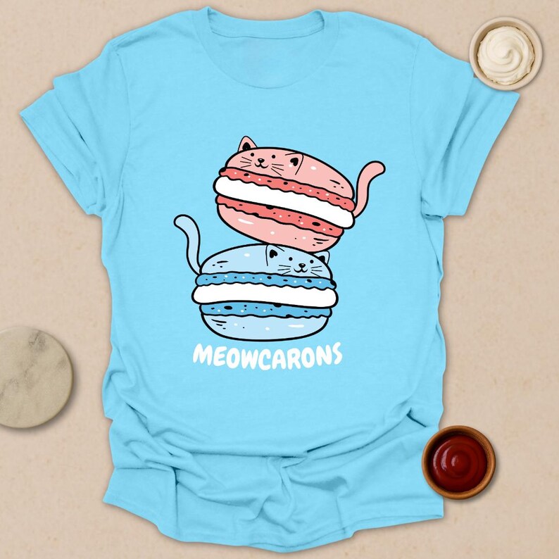 May include: Light blue t-shirt featuring a cartoon illustration of two cat-shaped macarons, one pink and one blue, stacked on top of each other. The text "MEOWCARONS" is printed below the macarons. The shirt is a casual wear item.