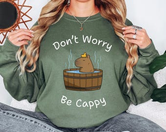 May include: A green sweatshirt with the text "Don't Worry Be Cappy" and a cartoon image of a capybara wearing a yellow rubber ducky in a wooden hot tub.