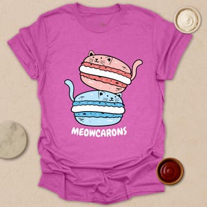 May include: A magenta t-shirt featuring a cartoon design of two stacked macarons, one pink and one blue, each with a cat face. The text "MEOWCARONS" is printed below the macarons in white.