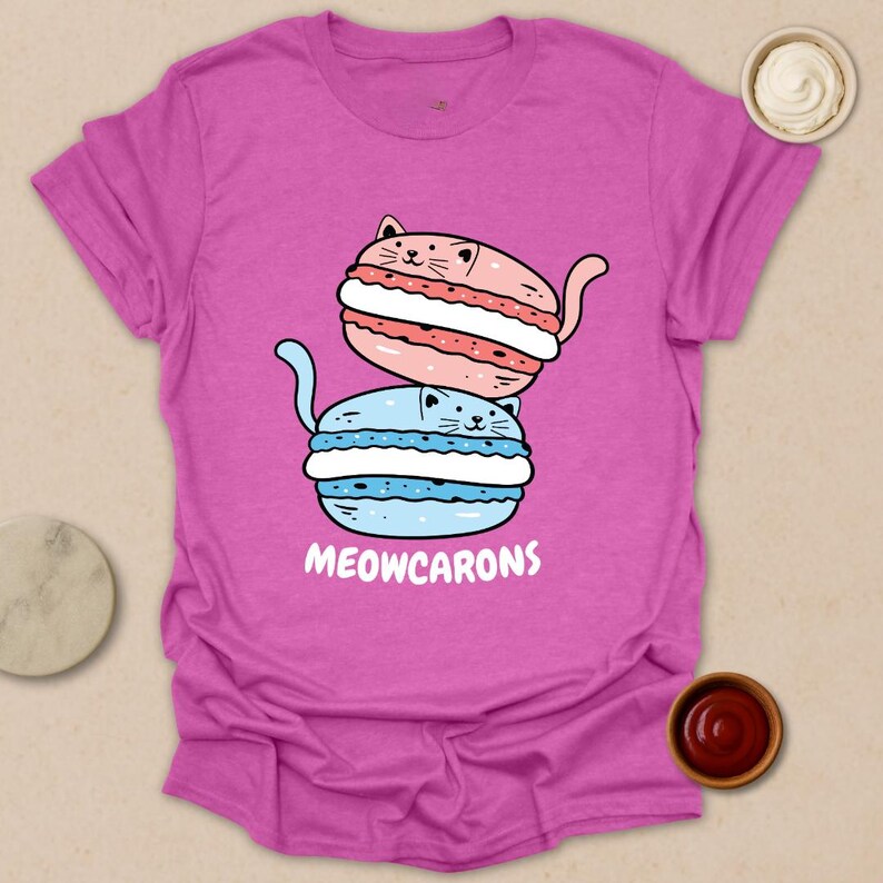 May include: A magenta t-shirt featuring a cartoon design of two stacked macarons, one pink and one blue, each with a cat face. The text "MEOWCARONS" is printed below the macarons in white.