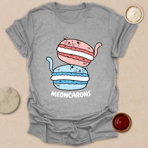 May include: A gray t-shirt featuring a whimsical design of two stacked cat-shaped macarons, one pink and one blue, with the word "MEOWCARONS" in white. The macarons have a cream filling and cat-like features.