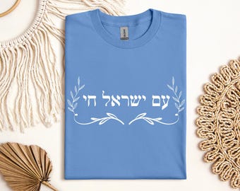 May include: A light blue t-shirt with the Hebrew text "עם ישראל חי" in white, meaning "The people of Israel live". The text is surrounded by stylized white floral designs.