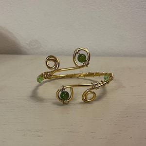 May include: Gold-toned wire arm cuff with green beads. The cuff features a swirling design with three larger green beads and smaller green beads. The jewelry is displayed on a light-colored surface.