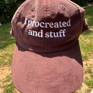 May include: A brown corduroy baseball cap with the embroidered text "I procreated and stuff" in white. The cap has a curved brim and a button on top. The background shows green grass and trees.