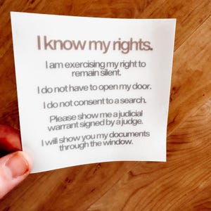May include: A white sticky note with the text "I know my rights" and a list of rights. The note is held by a person's fingers against a wooden background. The text is in a simple, easy-to-read font.