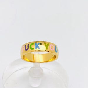 May include: Gold-toned ring with the words "FUCK YOU" in colorful letters. The ring has a smooth, polished surface and a bold, eye-catching design. The letters are in purple, blue, yellow, pink, and green.