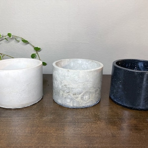 Op de afbeelding: Drie betonnen potten, een witte, een lichtgrijze en een zwarte. De potten zijn allemaal cilindrisch en hebben een glad, licht getextureerd oppervlak.