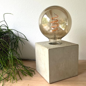 May include: A concrete lamp base with a clear glass light bulb with a coiled filament. The base is a cube shape and is a light gray color.