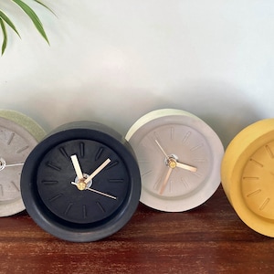Può includere: Quattro orologi in cemento di diversi colori: grigio, nero, beige e giallo. Ogni orologio ha un design semplice con lancette nere e un bordo rialzato.