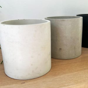 Op de afbeelding: Drie cilindrische betonnen plantenbakken, een wit, een lichtgrijs en een zwart. De bakken staan op een houten oppervlak.