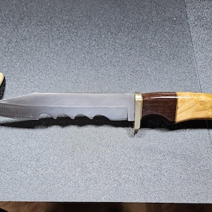 May include: A hunting knife with a serrated blade and a wooden handle. The handle is a combination of dark brown and light-colored wood, with a brass bolster. A small piece of bone rests near the blade.
