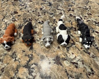 May include: Five small, detailed, realistic-looking dog figurines. The dogs are lying down and are made of a hard, smooth material. The dogs are brown and white, black and white, gray and white, black and white, and black and white.