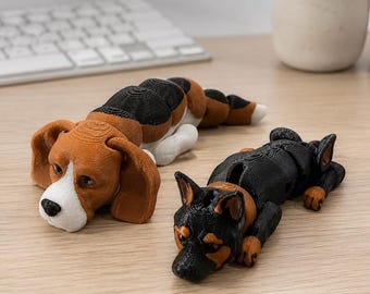 May include: Two articulated 3D-printed dog figurines. One is a beagle with brown, black, and white coloring, and the other is a black and tan dog. Both figures feature segmented bodies that allow for flexible movement and are positioned lying down.