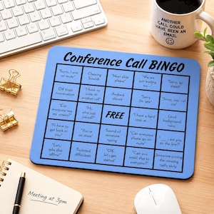 May include: A blue Conference Call Bingo game mat with phrases like "Sorry, I was on mute" and "Can everyone see my screen?" is displayed. A white keyboard, a coffee mug, and a notepad with "Meeting at 3pm" are also visible.