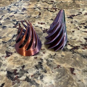 May include: Two 3D printed objects, one is a copper-colored cone with a spiral design and the other is a purple cone with a similar spiral design.