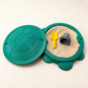 May include: A green turtle-shaped sandbox with a removable lid featuring a shell pattern. The open base contains beige sand, a small yellow plastic shovel, and a gray plastic bucket. The design includes molded details on the rim and lid.