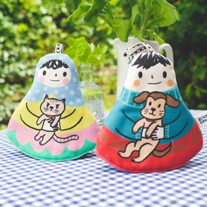 Op de afbeelding: Twee pluche poppen met cartoongezichten en dierenillustraties. De ene pop is lichtblauw met een kat, de andere is turquoise met een hond. Beide poppen hebben een lus aan de bovenkant om op te hangen. Op een blauw-wit geruit tafelkleed.