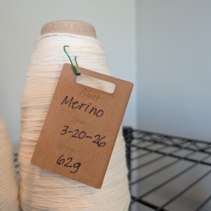 May include: A large cone of off-white yarn with a brown tag attached. The tag reads "Fiber: Merino, Spun: 3-20-26, grams: 62g". The yarn is tightly wound and has a cardboard core. A green wire secures the tag.