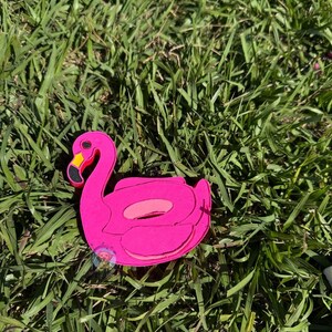 May include: A pink inflatable flamingo pool float with a yellow beak and black eyes. The float is lying on green grass.