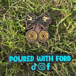 May include: A wooden cherry-shaped design with a camouflage bow and two 12-gauge bullet casings. Below, a black rectangle with the text "POURED WITH FORD" in turquoise, along with social media icons.