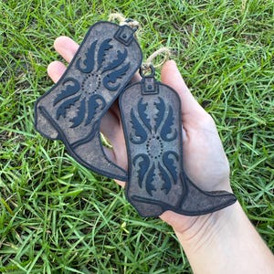 May include: Two wooden cowboy boot ornaments with black and brown paint and a floral design. The boots are hanging from twine.