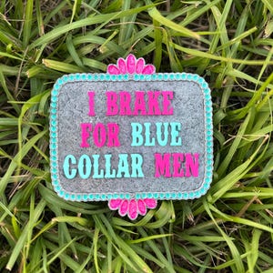 K&ouml;nnte beinhalten: Eine graue G&uuml;rtelschnalle aus Metall mit einem t&uuml;rkisfarbenen und rosafarbenen Rand. Die Schnalle tr&auml;gt den Text "I Brake For Blue Collar Men" in rosa und t&uuml;rkisfarbener Schrift.
