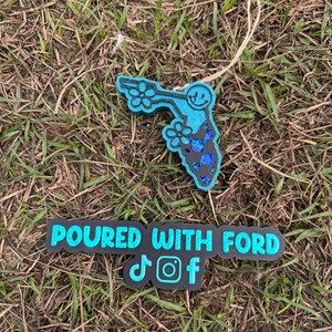 May include: A teal and black Florida-shaped ornament with a smiling face and flower details. The ornament is on a bed of grass. Below it is a black rectangle with the text "POURED WITH FORD" and social media icons.